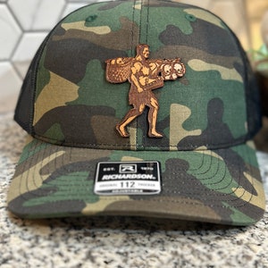 Op de afbeelding: Een camouflage trucker cap met een zwart mesh achterpaneel. De pet heeft een bruin leren patch met een man die een mand met schedels draagt. De patch heeft de tekst "Richardson" en "Est. 1970".