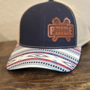 Op de afbeelding: Een marineblauwe en witte baseballpet met een bruin leren patch waarop "PRIDE" staat met gekruiste lacrosse-sticks achter het woord. De klep van de pet heeft een wit, blauw en rood geometrisch patroon.