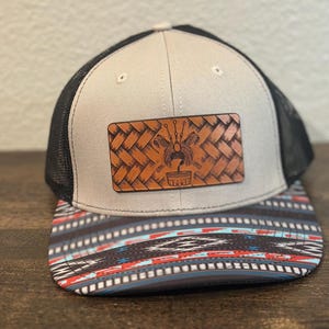 May include: A gray and black trucker hat with a brown leather patch featuring a woven design and a stylized image. The hat has a black mesh back and a colorful patterned brim.