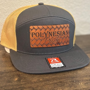 Op de afbeelding: Een grijze en beige flatbill snapback-pet met een bruin leren patch waarop "Polynesian Pride" in een geweven patroon staat.
