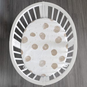 May include: An oval white crib with vertical slats containing a fitted sheet. The sheet is white and features a repeating pattern of light brown, circular, brushstroke-style spots interspersed with small, scattered light brown dots.