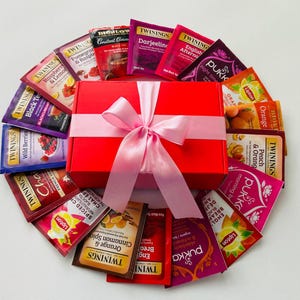 Puede incluir: Una caja de regalo roja atada con una cinta rosa, rodeada de varios sobres de té. Los sabores incluyen frambuesa, grosella negra, naranja y especias chai. Las marcas incluyen Twinings, Lipton y Pukka. Los sobres de té están dispuestos en círculo.