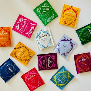 Può includere: Assortimento di bustine di tè alle erbe Pukka in vari colori, tra cui arancione, rosa, verde e giallo. Ogni bustina presenta il logo Pukka e nomi di gusti come "Three Ginger" e "Turmeric Active". Le bustine sono disposte su una superficie bianca.