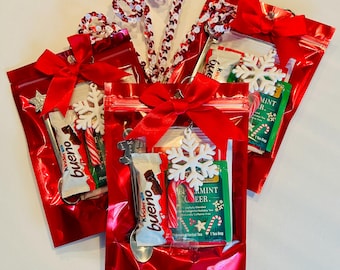 May include: Red cellophane gift bags tied with red bows, each containing a Kinder Bueno chocolate bar, peppermint candy, snowflake ornament, and tea bag. Decorative candy cane spoons and star accents complete the festive holiday theme.