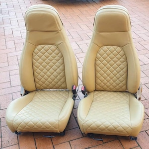 May include: A pair of tan leather car seats featuring diamond-stitched detailing. Each seat has an integrated headrest and seatbelt. The seats are designed for a vehicle and are in a neutral colour.