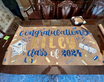 May include: A brown paper banner with the words "Congratulations Riley Class of 2024" written in blue and gold paint. The banner is decorated with gold and blue confetti, a graduation cap, and a diploma.