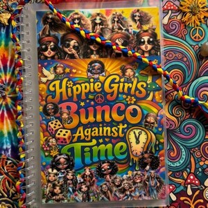 May include: A spiral-bound notebook with the title "Hippie Girls Bunco Against Time" in a colorful, psychedelic design. The cover features cartoon illustrations of women. A red pouch with dice and a braided cord are also visible.