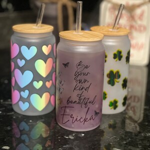 May include: Three glass tumblers with bamboo lids and clear straws. One tumbler has a pattern of rainbow hearts, another has the text "Be your own kind beautiful Ericka", and the third has green shamrocks.
