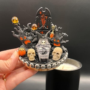 May include: A Halloween-themed candle lid featuring a miniature spooky scene. The lid includes a coffin, skulls, and a throne, with orange and black accents. The candle itself is in a black container.