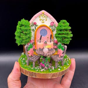 May include: A miniature fairy house scene featuring a pink arched door, two green trees, and a small round table with two cups of bubble tea. The base is covered in green grass, pink flowers, and two brown chairs, all set on a circular wooden slice.