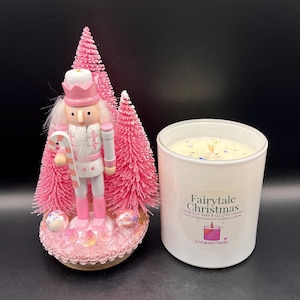 May include: A nutcracker figurine in pink and white attire holding a candy cane, positioned in front of three pink bottle brush trees on a decorated base, next to a white candle jar labeled Fairytale Christmas 100% Soy Wax 8 oz/292 grams.
