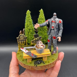 May include: A diorama featuring a headless zombie figure in a grey suit with red blood stains, standing near two green pine trees and a tombstone marked RIP. A severed head sits in a small grey pot on a wooden bench with a bloody knife and a black spider.