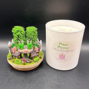 May include: A white candle in a cylindrical jar with a label reading Pixie Picnic 100% Soy Wax 8 oz/292 grams. Beside it is a miniature garden scene featuring two brown chairs, a round table with two green drinks, and two green trees.