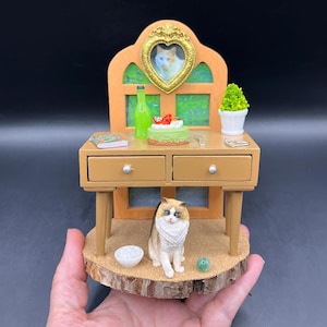 May include: A miniature wooden desk with two drawers, topped with a green bottle, a small cake, a book, a fork, a heart-shaped frame with a cat photo, and a potted plant. A cat figurine sits below on a wood slice base with a bowl and a green marble.