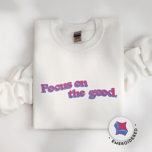 Peut inclure: Sweat-shirt blanc avec le texte "Focus on the good." brodé en lettres roses et bleues.