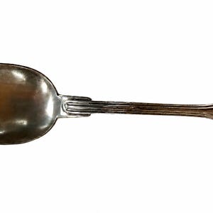 May include: A silver-toned metal spoon featuring a polished oval bowl and a slender handle with vertical ribbed detailing near the neck. The handle end is rounded with a decorative border and features the engraved cursive script text Paubi.
