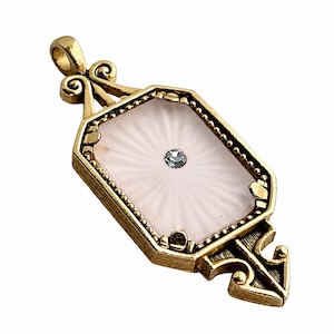 May include: A gold-toned metal pendant featuring an octagonal frame with decorative scrollwork at the top and bottom. The center contains a pale pink, sunburst-patterned inlay with a single small, clear, round gemstone set in the middle.
