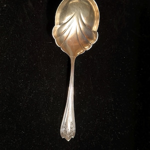 May include: A silver serving spoon with a decorative, leaf-like design on the bowl. The handle tapers to a point and features an ornate floral pattern. The spoon is set against a dark background.