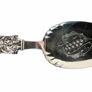May include: A silver-toned fish serving utensil featuring an ornate, embossed handle with intricate scrollwork and a wide, oval-shaped blade. The blade displays a central engraved design of two fish facing opposite directions with a grid pattern.