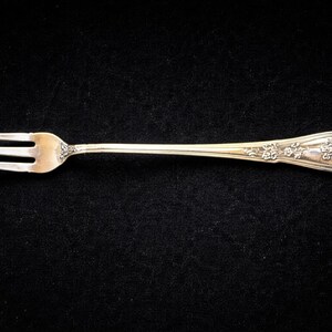 May include: A silver fork with three tines and a floral design on the handle. The fork is set against a black background, highlighting its intricate details and elegant form. This utensil is likely for serving appetizers or desserts.