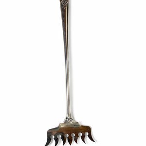 May include: A silver-colored serving fork with a long, slender handle and a decorative floral design at the top. The fork has a wide, flat head with multiple tines, ideal for serving. The utensil is likely vintage or antique.