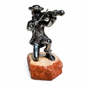 May include: A polished silver-toned figurine of a musician kneeling while playing a violin. The figure wears a hat and coat and is mounted on a rough-hewn, rectangular reddish-brown stone base with a small white label featuring black text.