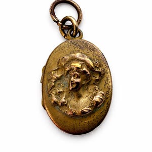 May include: Oval-shaped antique brass locket with a detailed relief of a woman's face. The locket has a loop at the top for a chain and shows signs of age and wear, giving it a vintage aesthetic. The woman's face is framed by ornate details.