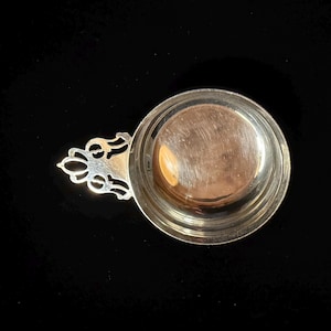 May include: A small, silver-colored cup with a decorative handle. The cup has a round bowl and a detailed, ornate handle. The handle features a floral design. The cup is set against a black background.