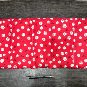 May include: A rectangular red pet bed with a white paw print and heart pattern. The bed is divided into sections and is on a dark wooden surface. A pen is below the bed.