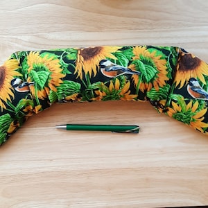 May include: A long, curved neck pillow with a sunflower and bird print. The pillow is made of fabric and has a black background with yellow sunflowers and small birds.