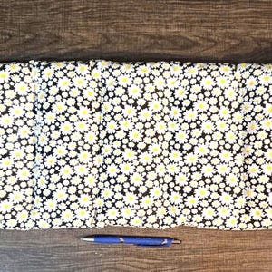May include: Rectangular pillow with a black background covered in white daisies with yellow centers. A blue pen rests on the wooden surface below the pillow. The pillow is likely for home decor.