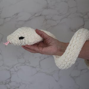 May include: A plush, off-white snake toy with a textured body and a pink tongue. The snake's head features black eyes. The toy is held in a hand against a marble-patterned background. The snake is a soft, cuddly toy.