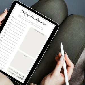 May include: A digital tablet displaying a daily goals and priorities planner with sections for hourly planning, notes, weather, and mood. The tablet is on a person's lap and a white stylus is being held in their hand.