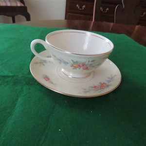 May include: A cream-colored teacup and saucer set with delicate floral patterns in pink, yellow, and blue. The cup has a curved handle and a gold rim, resting on a matching saucer. The set is placed on a green surface.