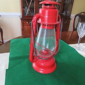 May include: A red vintage kerosene lantern with a clear glass globe and a black handle. The lantern is sitting on a green tablecloth.