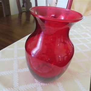 May include: A vibrant red glass vase with a flared top and a rounded body. The vase is made of transparent glass, allowing light to pass through. It sits on a cream-colored tablecloth with a geometric pattern. The vase is suitable for floral arrangements.