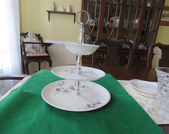May include: Two-tiered serving stand with a silver base and white ceramic plates. The plates have a floral design with pink flowers and green leaves.