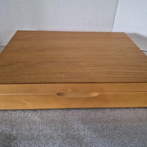 May include: A rectangular wooden storage box with a smooth, light brown finish. The box features a flat, grain-patterned lid and a front panel with a centered, horizontal oval cutout designed to serve as a handle for opening the container.
