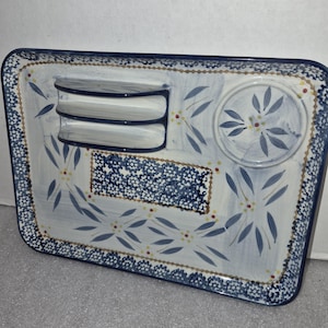 Puede incluir: Una bandeja de servicio rectangular de cerámica con base blanca, patrones florales azules, detalles amarillos y un borde azul oscuro. La bandeja incluye un soporte integrado de dos niveles, una hendidura circular y una sección rectangular texturizada.
