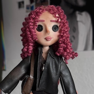 May include: A doll with curly magenta yarn hair, button eyes, and a small nose ring. The doll wears a black faux leather jacket over a dark shirt, dark trousers, and carries a small brown shoulder bag. The facial features include pink eyebrows.
