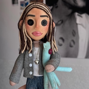 May include: A handmade doll with button eyes, long brown hair with blonde highlights, and pink lips. The doll wears a grey herringbone jacket over a white shirt, blue jeans, and a silver heart necklace, while holding a light blue fabric cat toy.
