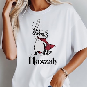 May include: White t-shirt featuring a cartoon raccoon wearing a red cape and holding a sword aloft. The raccoon has a black mask and is standing on its hind legs. The word "Huzzah" is printed in black script below the raccoon.