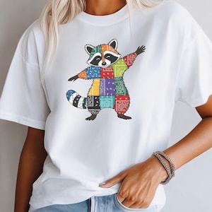 May include: White t-shirt featuring a colorful raccoon graphic. The raccoon is designed with a patchwork pattern of various colors and bandana prints, with its arms outstretched in a dabbing pose. The shirt is a casual, everyday wear item.