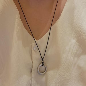 May include: A silver pendant necklace with a black cord. The pendant is a rounded, open circle. The necklace is shown against a cream-coloured, button-down shirt.