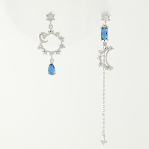 Peut inclure: Une paire de boucles d'oreilles en argent avec un motif de croissant de lune et d'étoile. Une boucle d'oreille a une pierre précieuse bleue en forme de larme qui pend, tandis que l'autre boucle d'oreille a une chaîne plus longue avec un charme d'étoile à l'extrémité.