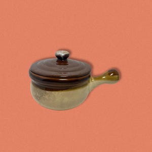 Può includere: Una pentola per zuppa in ceramica marrone e beige con un coperchio e un manico. La pentola ha una glassa macchiata.