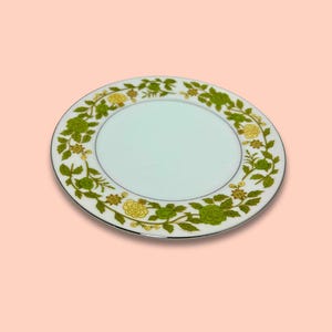 Könnte beinhalten: Weiße Keramikplatte mit floralem Design. Der Rand hat ein grünes und gelbes Blumenmuster mit Blättern. Der Teller hat einen silbernen Rand und eine hellblaue Mitte.