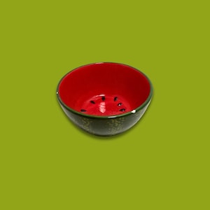 May include: A ceramic bowl with a watermelon design. The interior is red with black seed details, and the exterior is green. The bowl is set against a green background, creating a vibrant contrast. This bowl is ideal for serving snacks or small portions.
