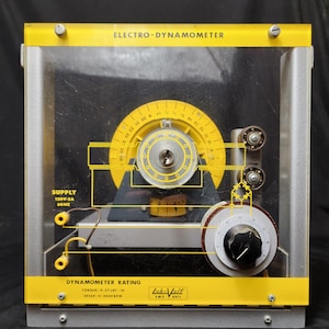 May include: An electro-dynamometer with a yellow and silver frame. The device has a clear front panel, a circular dial, and a yellow semi-circular scale. Text on the device includes "ELECTRO-DYNAMOMETER" and "DYNAMOMETER RATING".
