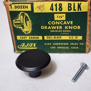 May include: A dull black concave drawer knob with a beveled edge, made of cast zamak, accompanied by a silver metal screw. The knob is displayed in front of its original green and yellow cardboard box labeled 418 BLK, 1 1/2 inch, Ajax Hardware.